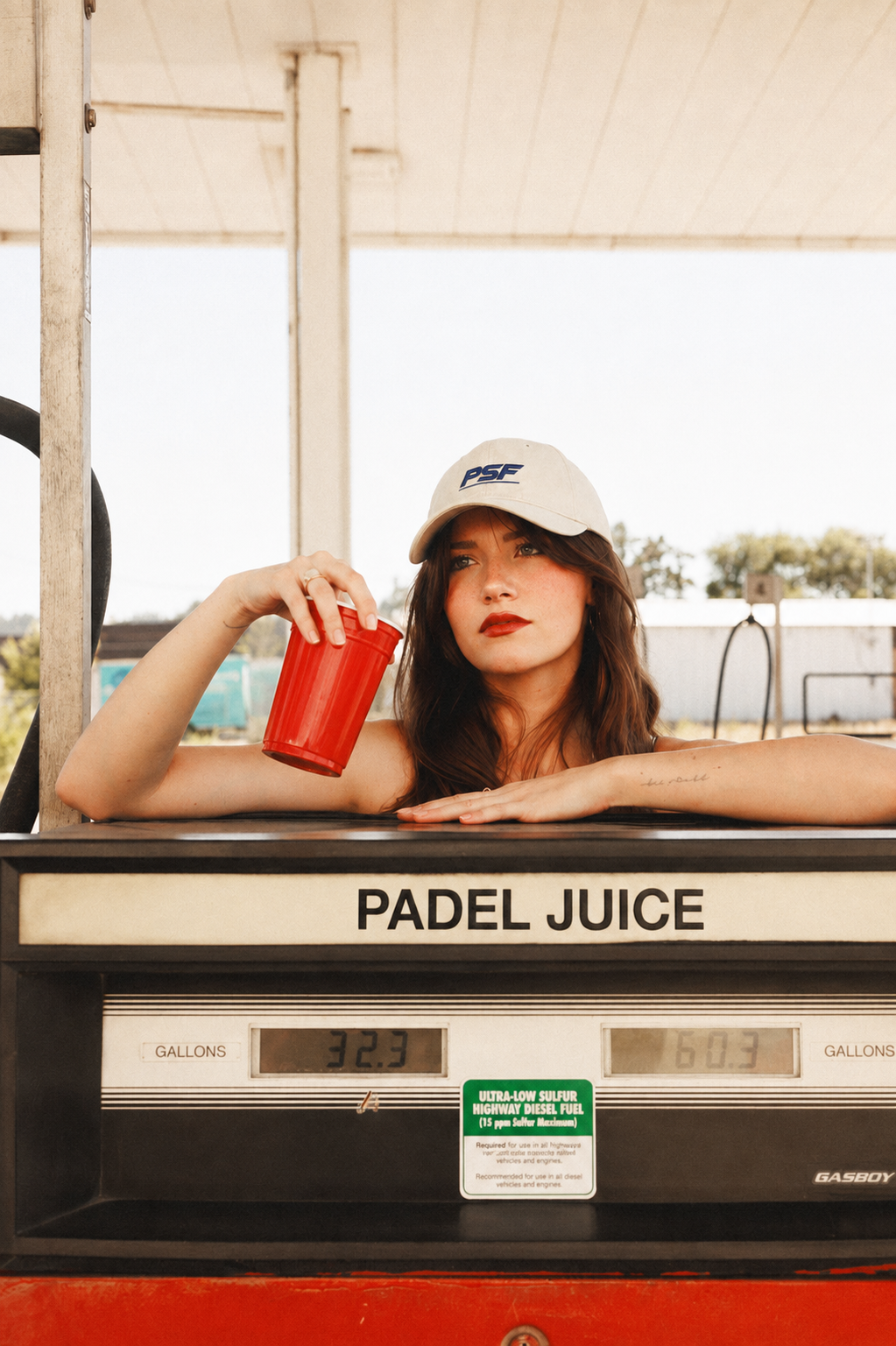 A woman in a white cap holds a red cup, leaning on a gas pump labeled PADEL JUICE. She looks at the camera, with sunlight and blurred trees in the background. Total: 174 characters.