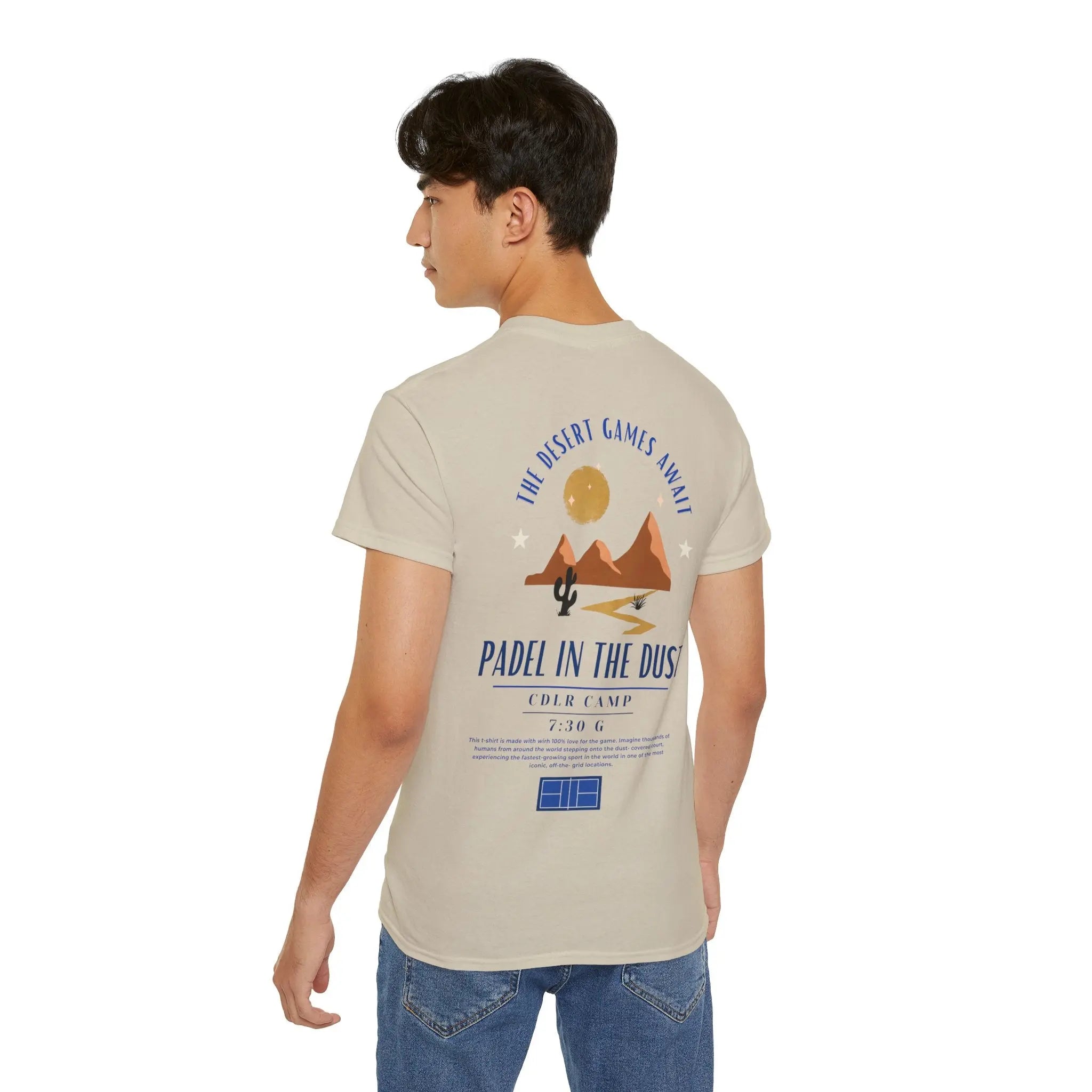 PSF Collective A young man faces away, wearing the Limited Edition - Padel in the Dust Tee by Printify. The back features desert mountains, a cactus, sun, and “The Desert Games Await—Padel in the Dust.” Ideal for Patty Camps or Burning Man padel events. Designed for the Unwind