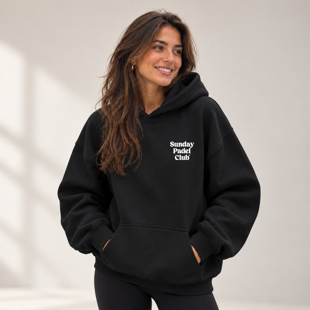 PSF Collective A smiling woman with long brown hair wears an oversized black Sunday Padel Club Hoodie with her hands tucked into the front kangaroo pocket, standing against a plain, light-colored background. Designed for the Unwind