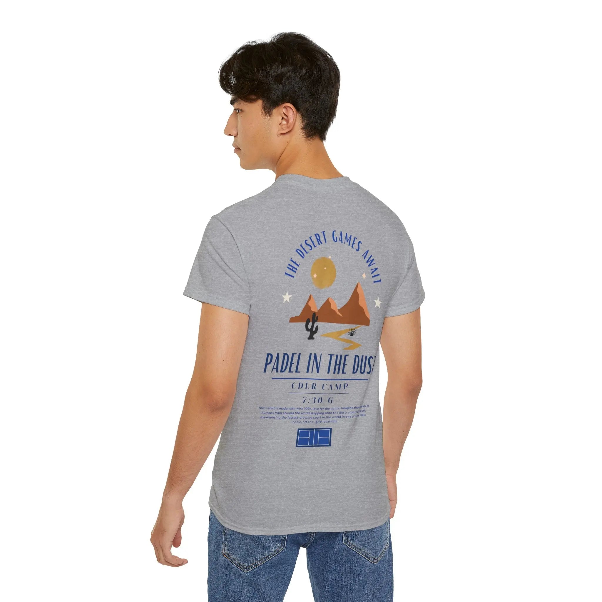 PSF Collective A young man with short dark hair wears the Printify Limited Edition - Padel in the Dust Tee, which has a desert graphic and text on the back, with blue jeans, posing against a plain white background. Designed for the Unwind