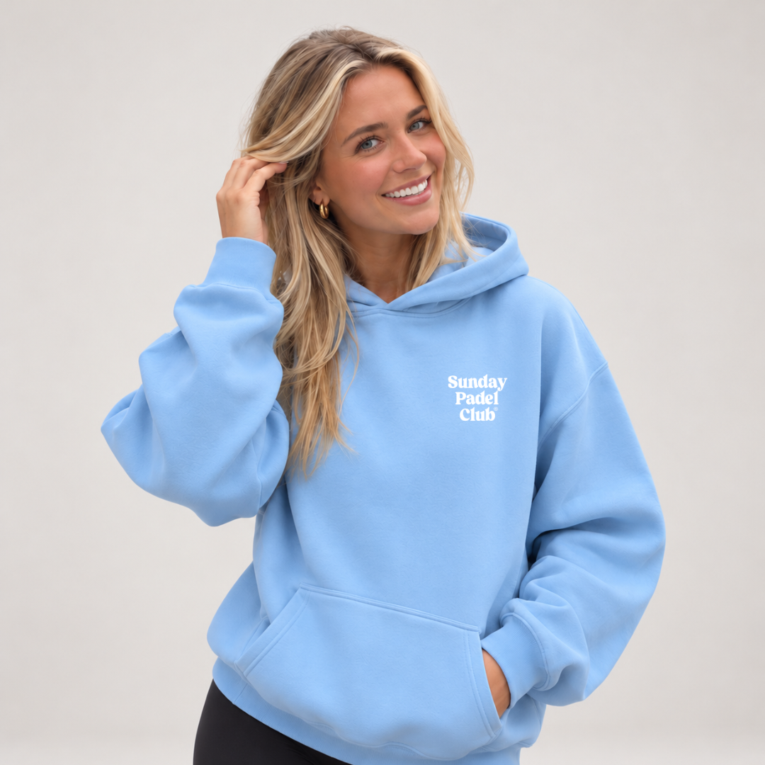 PSF Collective A smiling blonde woman wears a light blue Sunday Padel Club Hoodie in Carolina Blue with the name printed in white on the chest, her hand tucked into the front pocket, posed against a plain, light grey background. Designed for the Unwind