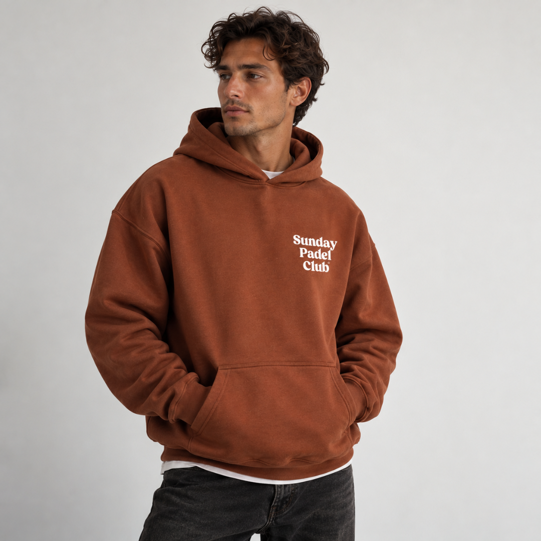 PSF Collective A man with wavy dark hair wears a clay-colored Sunday Padel Club Hoodie with the text printed in white on the chest, posing against a plain, light gray background with his hands tucked into the front kangaroo pocket. Designed for the Unwind