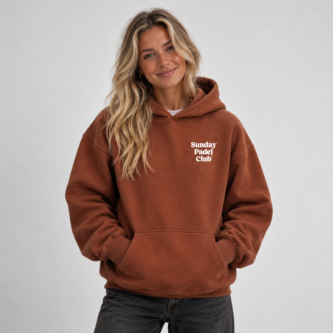 PSF Collective A woman with long blonde hair smiles wearing a clay-colored Sunday Padel Club Hoodie with a front kangaroo pocket and dark gray jeans against a light gray background. Designed for the Unwind