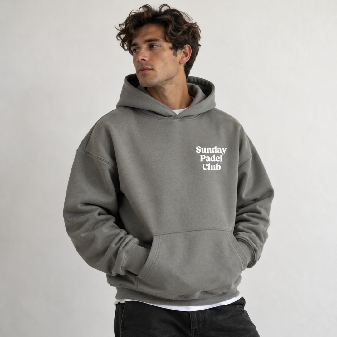 PSF Collective A man with wavy brown hair looks away from the camera, wearing an oversized gray Sunday Padel Club Hoodie in Shadow over a white shirt and dark jeans against a plain off-white background. Designed for the Unwind