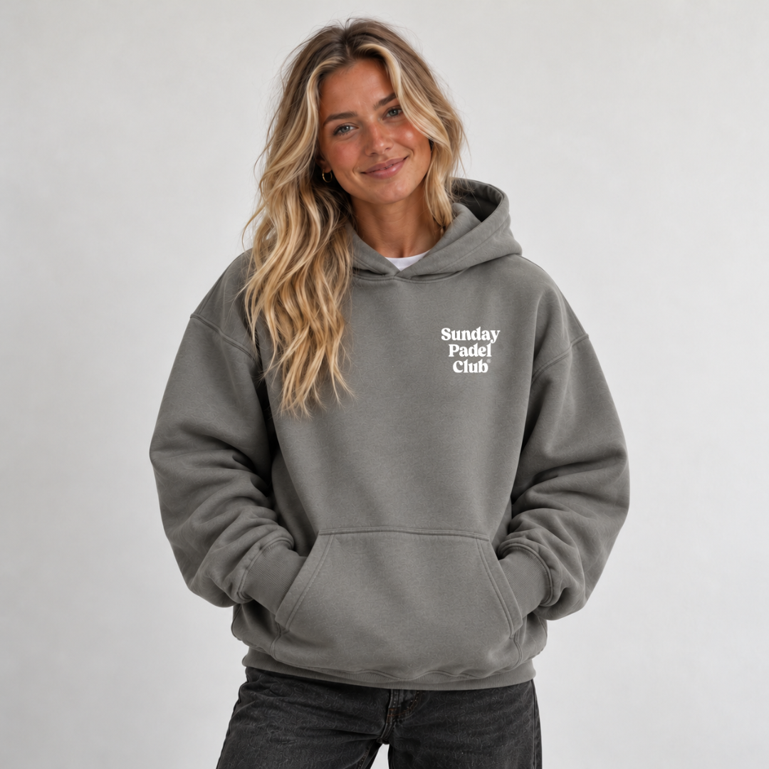 PSF Collective A smiling woman with long blonde hair wears an oversized, charcoal-gray Sunday Padel Club Hoodie in Shadow, standing against a light gray background with her hands tucked into the front kangaroo pocket to embody a relaxed padel lifestyle. Designed for the Unwind