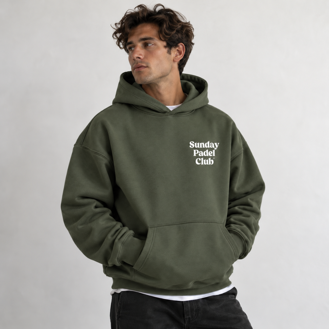 PSF Collective A man with wavy brown hair looks away from the camera wearing an olive green Sunday Padel Club Hoodie in Cypress. The sustainable hoodie features a front kangaroo pocket over a white t-shirt, paired with dark pants against a plain gray background. Designed for the Unwind
