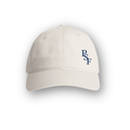 PSF Collective The Heritage Cap in Off White features a small dark blue PSF logo embroidered on the front left panel. Visible stitching and a curved brim evoke a classic vintage sportswear style. The cap is photographed against a white background with a soft shadow beneath it. Designed for the Unwind