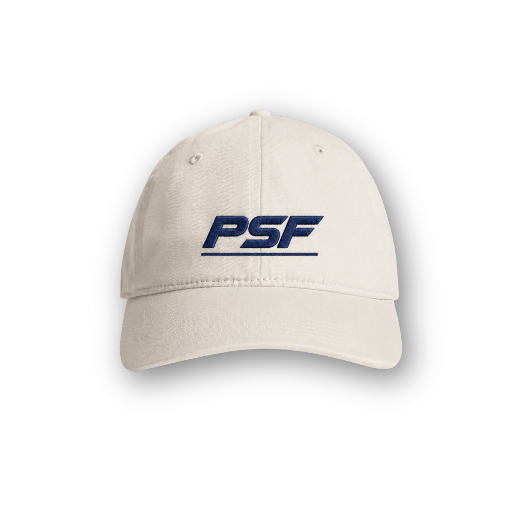 PSF Collective The Retro Cap is a beige hat with PSF embroidered in bold, dark blue letters on the front. It features visible stitching along the brim and two small ventilation holes on each side, capturing the essence of classic 80s sportswear. The cap is showcased against a plain white background with a soft shadow beneath it. Designed for the Unwind